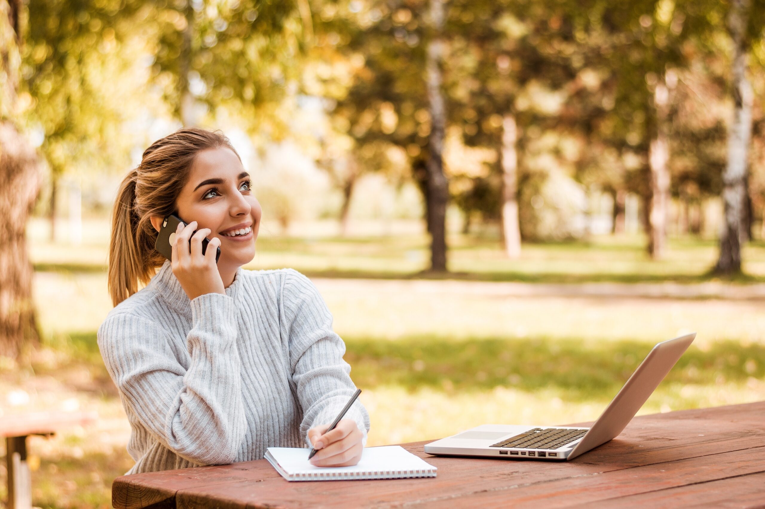 Young blonde woman talking on phone while taking notes outside.