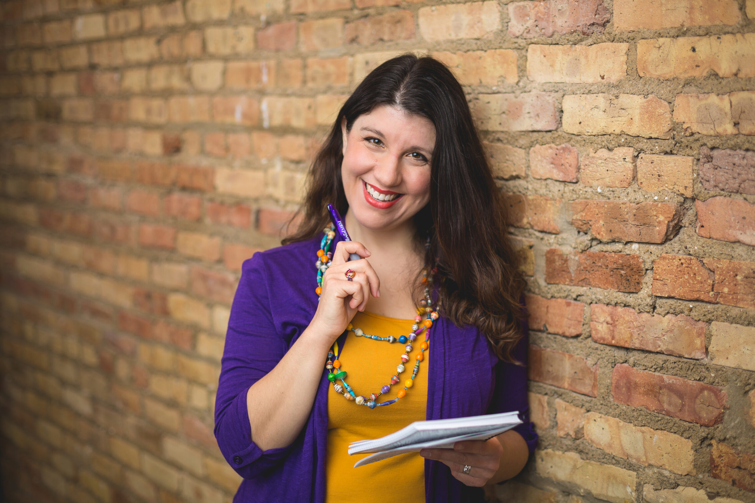 Valerie smiling at camera holding a pen in one hand and notebook in the other