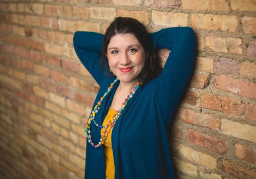 Valerie in teal and yellow leaning against a brick wall and smiling