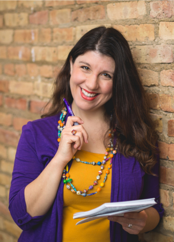 Valerie in purple and yellow holding a notebook and pen, leaning against a brick wall and smiling
