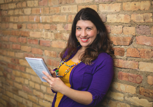 Valerie in yellow and purple leaning back against a brick wall holding a notebook and smiling